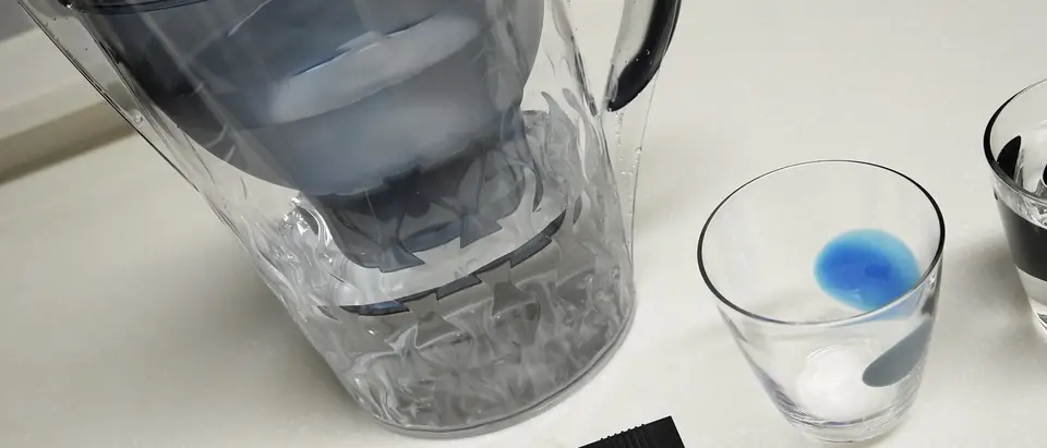 A water filter pitcher beside a glass of water and a test pen on a kitchen counter