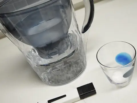 A water filter pitcher beside a glass of water and a test pen on a kitchen counter